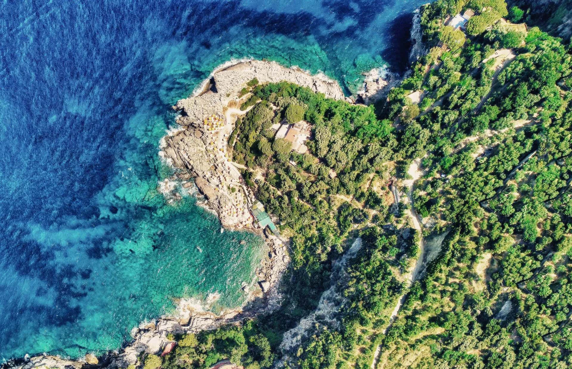 Aerial view of rocky coastline, dense green vegetation, and clear turquoise Mediterranean water at Punta Campanella.