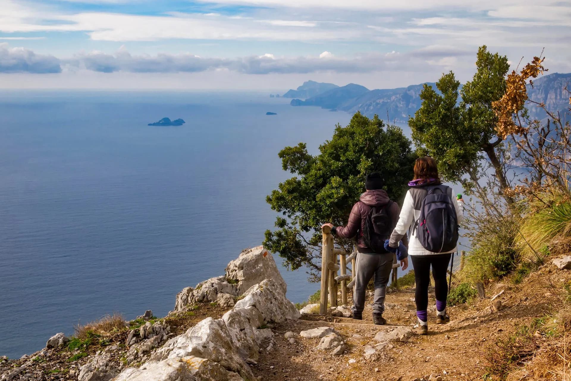 Hikers descending on a coastal path overlooking the deep blue sea and distant mountains.