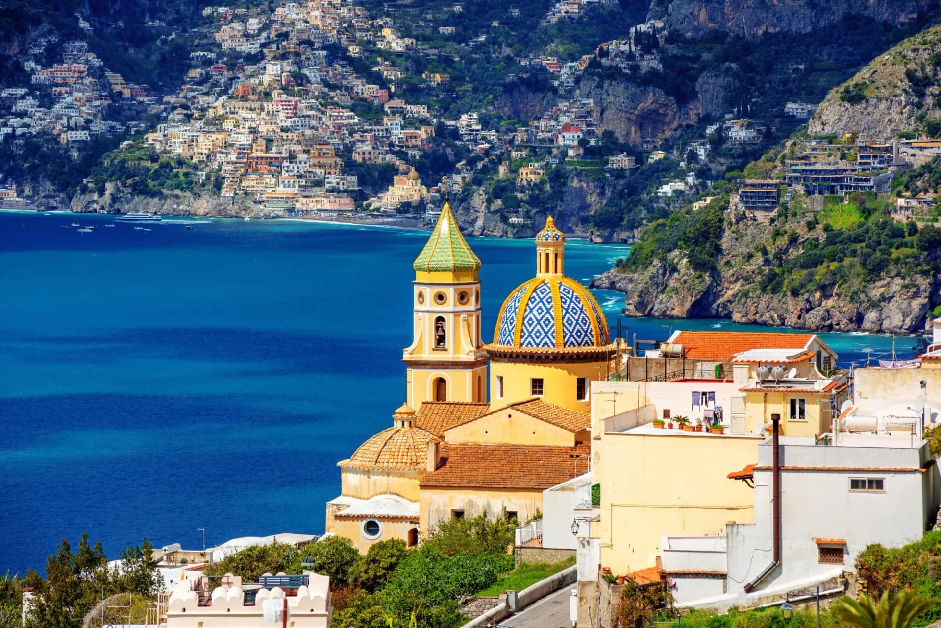 Church with tiled dome overlooking colorful town on steep cliffs above blue sea in Praiano.
