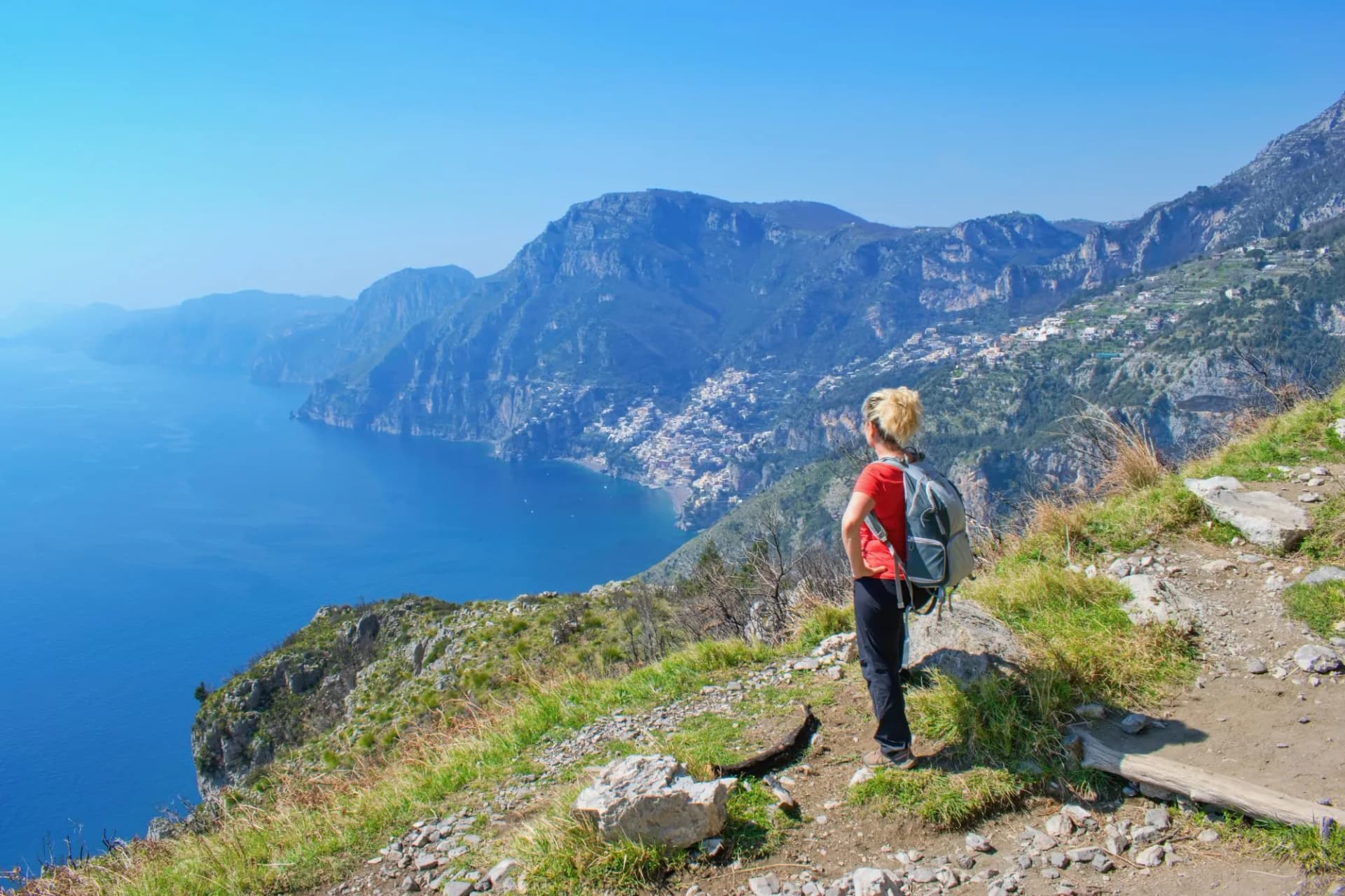 Hiker watching coastal scenery with steep mountains and a town overlooking the bright blue sea.