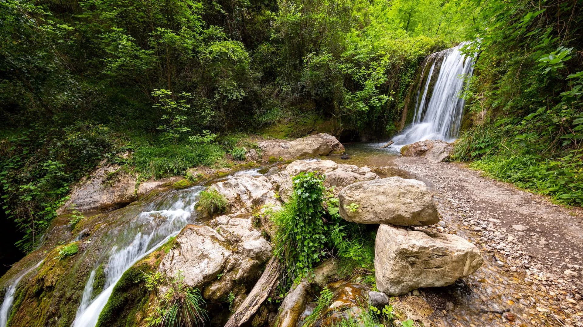 Waterfall cascading into a stream near a rocky hiking path in the Valley of the Ferriere.