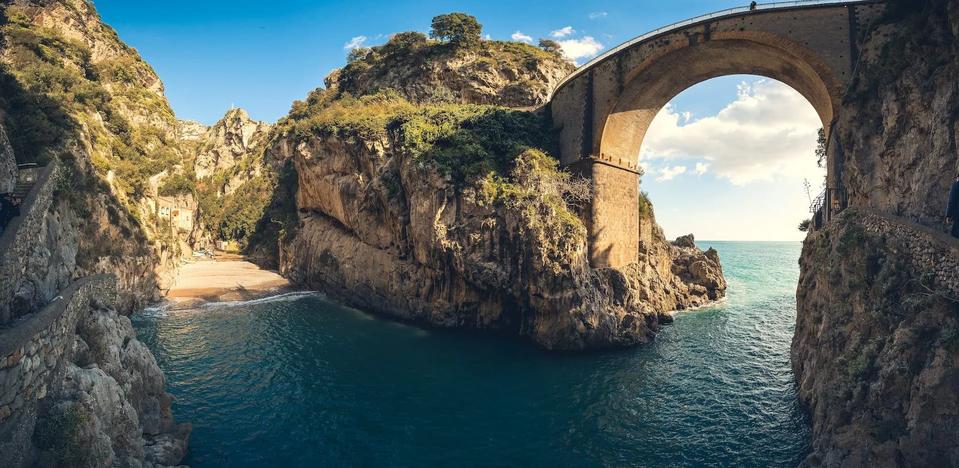 Stone arch bridge over turquoise sea inlet with small beach nestled between steep, rocky cliffs.