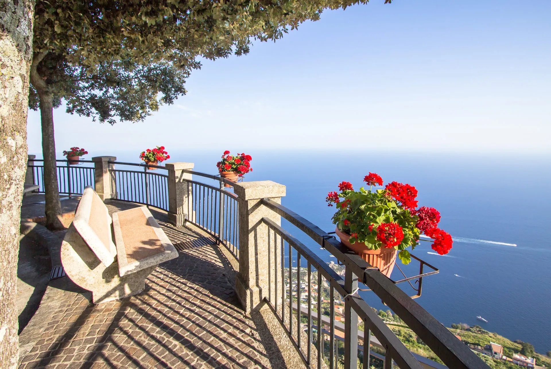 Scenic overlook with stone bench, red geraniums, and high view of blue sea and coastal village.