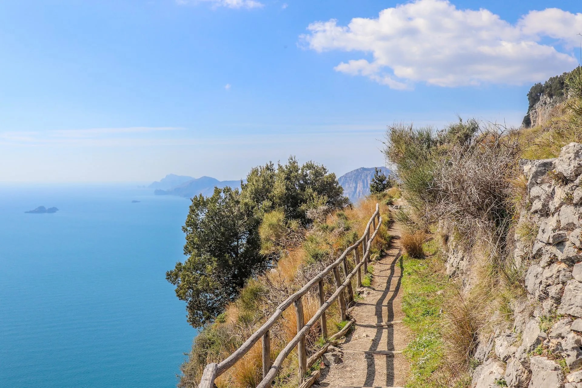 Hiking path with wooden railing along cliffs overlooking bright blue sea and distant mountains