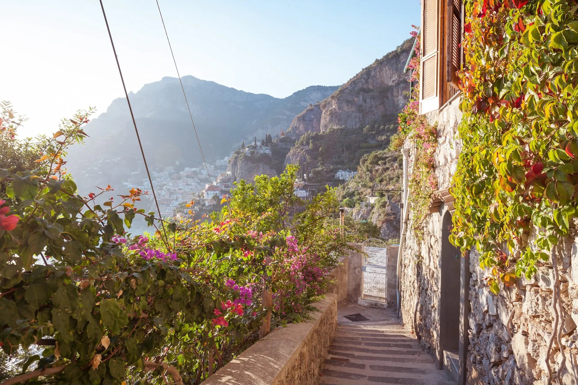 Stone staircase descending past flowering vines toward a coastal town nestled in mountains.