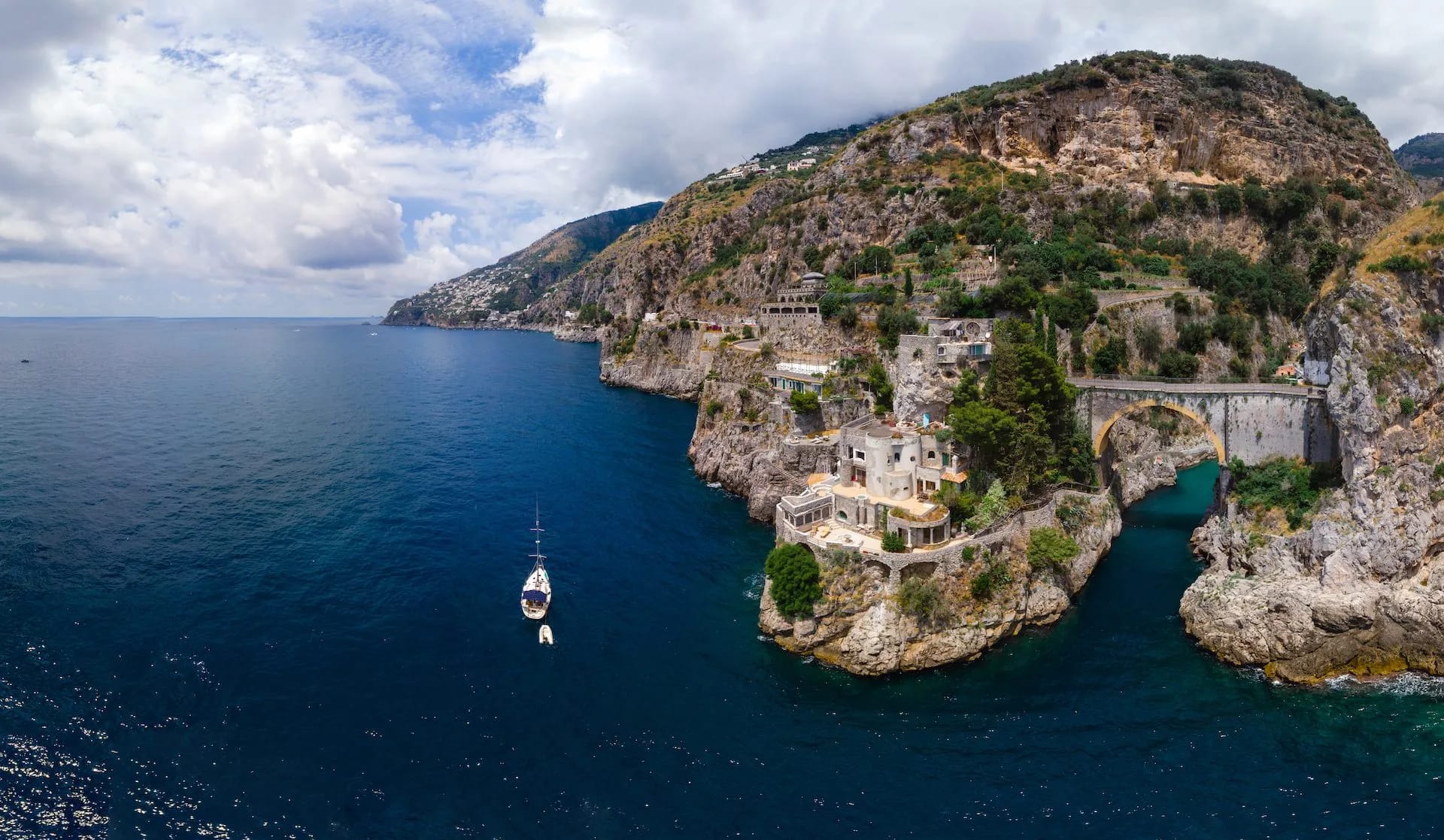 Sailboat near Furore Fjord with cliffside buildings and arched bridge on the Amalfi Coast.