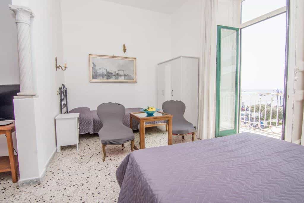 Hotel room with beds, chairs, and balcony view of the sea and coast.