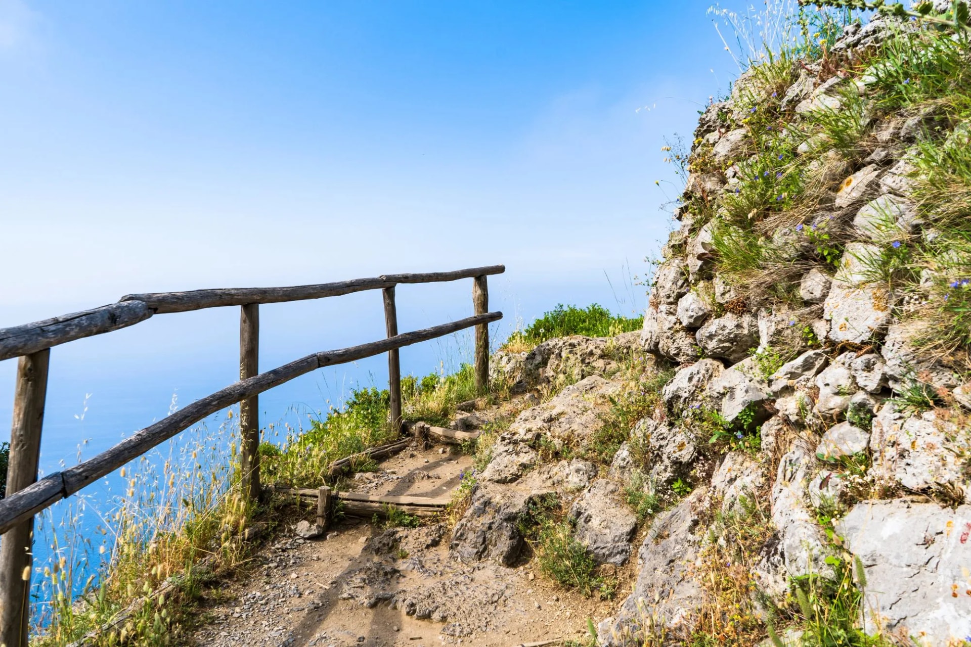 Hiking trail with wooden railing overlooking blue sea under clear sky